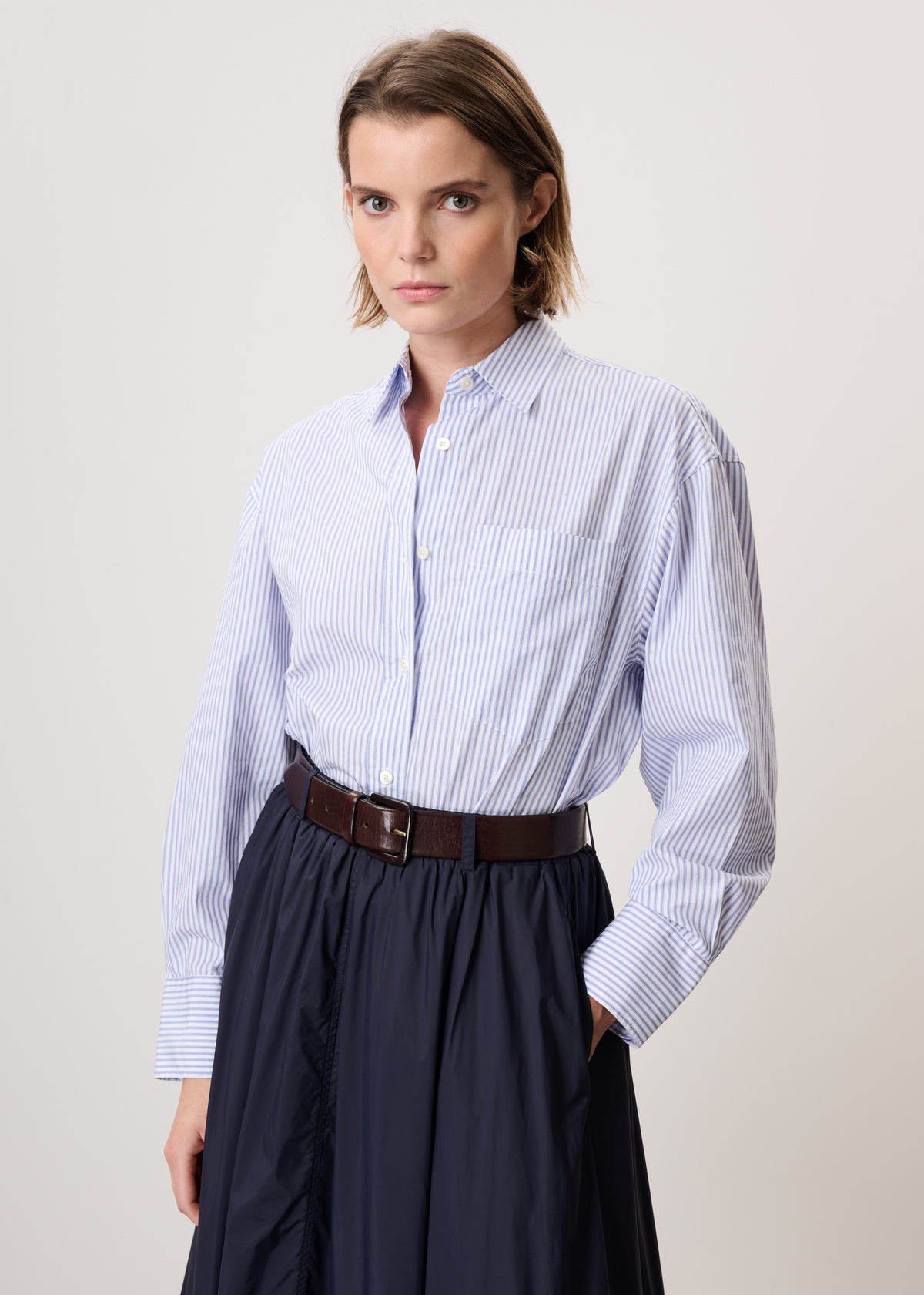 A woman modeling a light blue and white vertically striped button-down shirt tucked into a dark navy skirt, accessorized with a brown belt, against a plain light gray studio background.