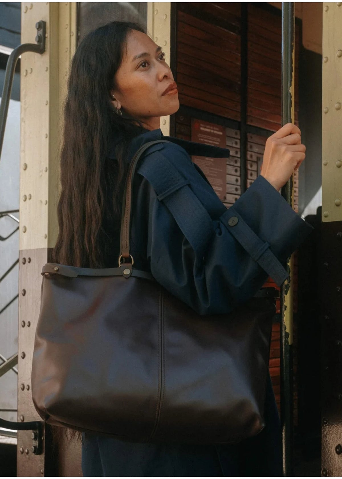 A woman wearing a navy blue jacket carries a large dark brown leather tote bag over her shoulder while standing in front of a vintage train car.