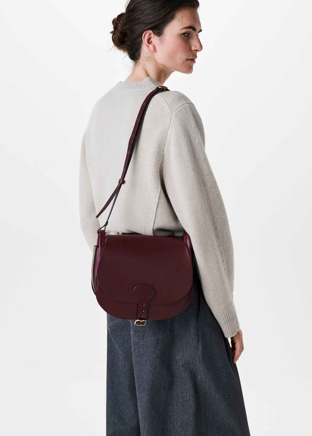 A woman wearing a light beige knit sweater and dark gray skirt, shown from a side-back angle while wearing a burgundy leather crossbody bag with gold hardware against a plain white background.