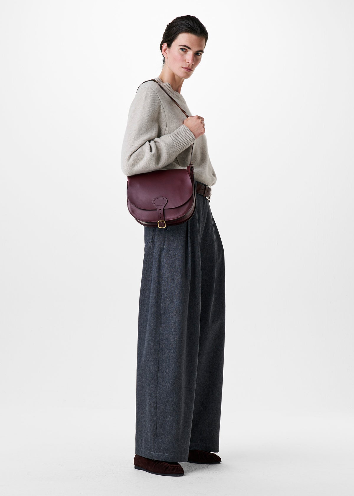 A model is shown from the waist up, wearing a light beige knit sweater, dark gray wide-leg trousers, and dark brown shoes, while carrying a burgundy leather shoulder bag. The background is a clean, neutral white studio backdrop.