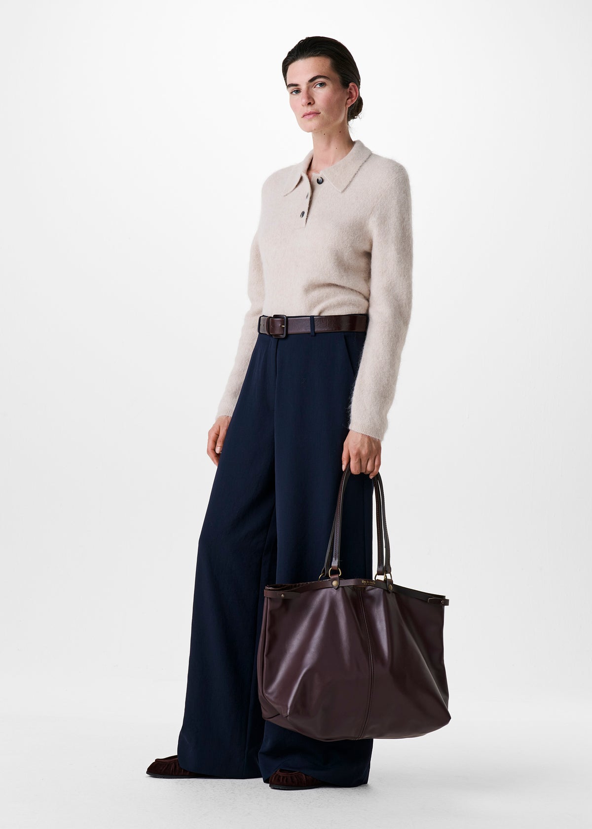 A woman stands against a light gray studio background wearing a cream knit polo sweater, dark navy wide-leg trousers, and holding a large dark brown leather tote bag.