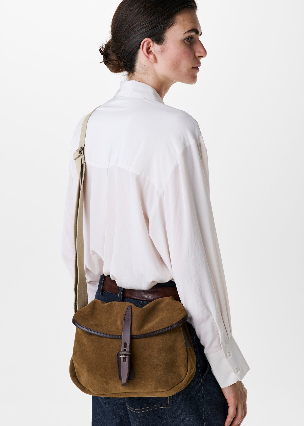 A woman in a white shirt and dark jeans is shown from the back and side, wearing a brown suede messenger bag with a dark brown leather strap and buckle, set against a plain white studio background.